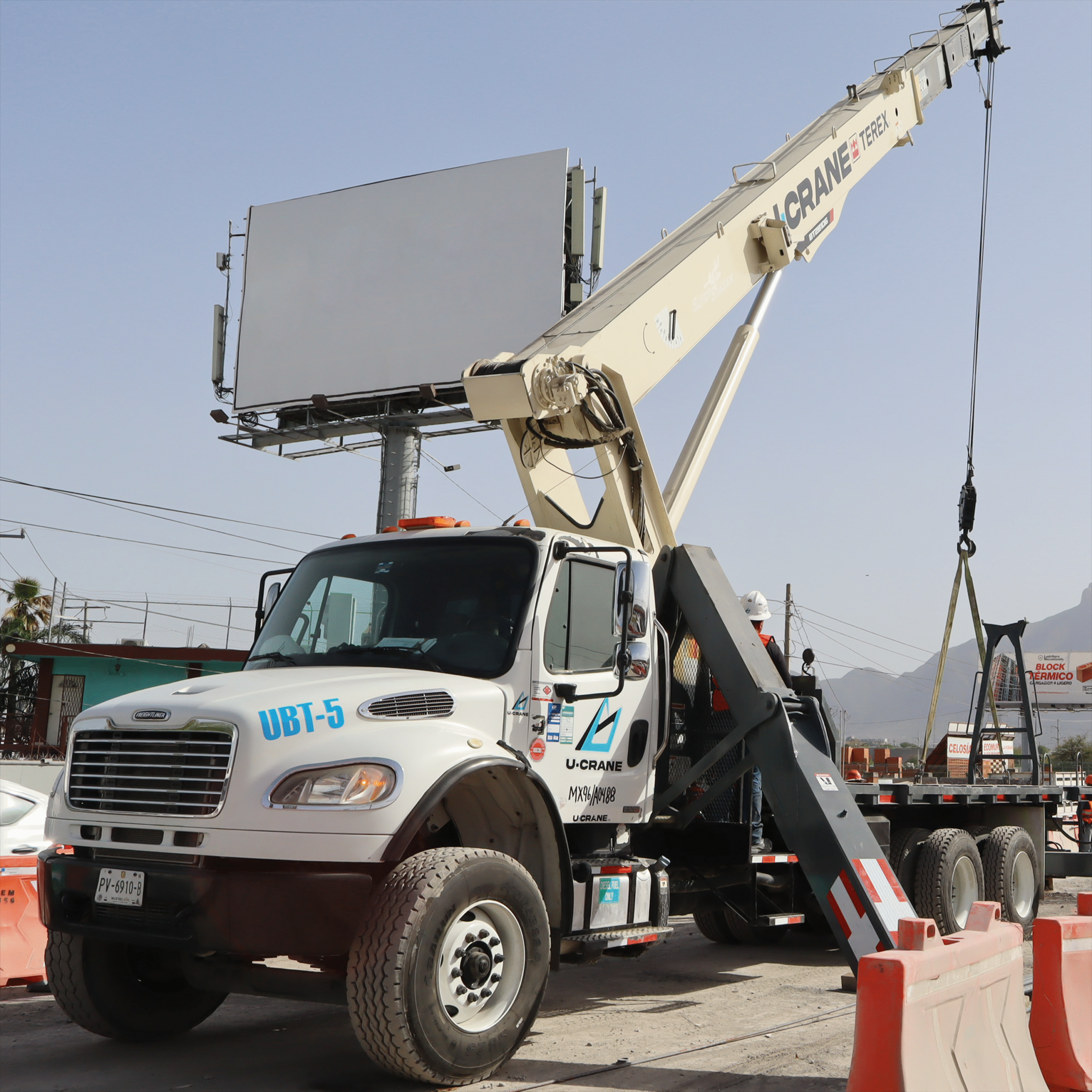 Grúa hidráulica Terex montada sobre camión Freightliner, operada por U-CRANE, realizando levantamiento de cargas en un entorno urbano. Esta maquinaria es adecuada para montaje de estructuras, instalación de anuncios publicitarios y manejo de cargas pesadas en espacios confinados. Imagen perfecta para temas relacionados con renta de grúas, logística urbana y servicios industriales en áreas metropolitanas.