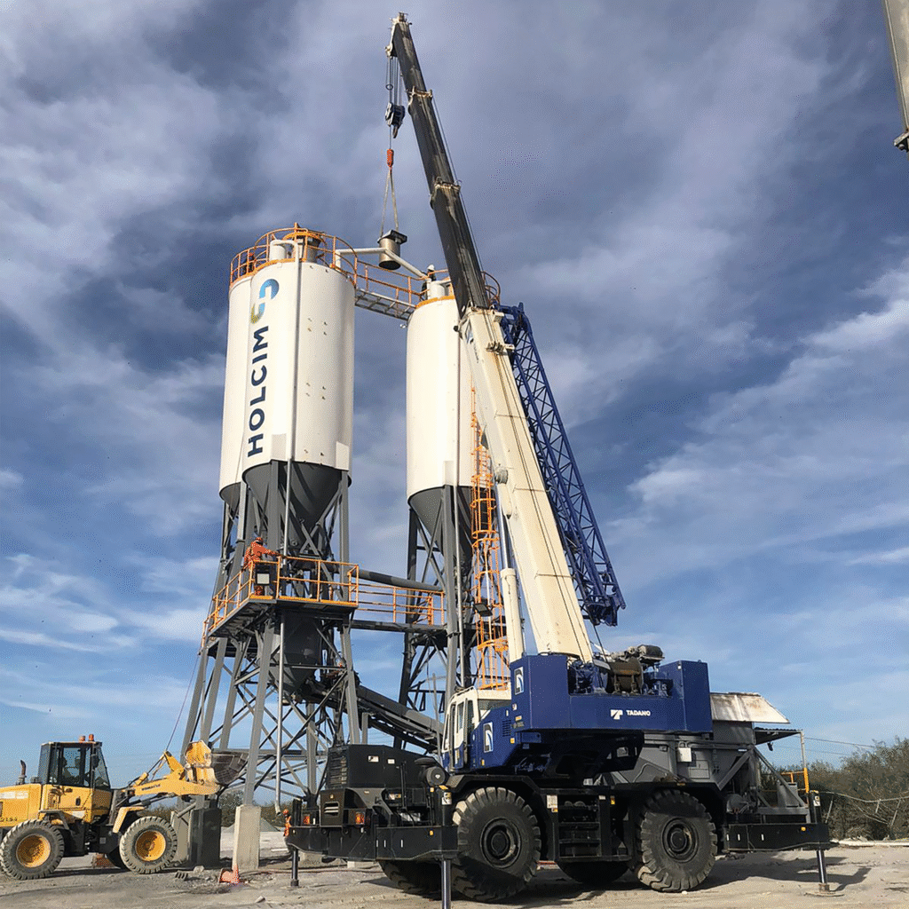 Montaje de silos de cemento en una planta industrial de HOLCIM, con una grúa hidráulica todoterreno Tadano elevando piezas para ensamblar la estructura superior del silo. La tarea se realiza con personal técnico capacitado y equipo pesado, garantizando seguridad en cada paso. Perfecto para ilustrar temas de construcción industrial, manejo logístico de materiales y uso de grúas en la industria del cemento.